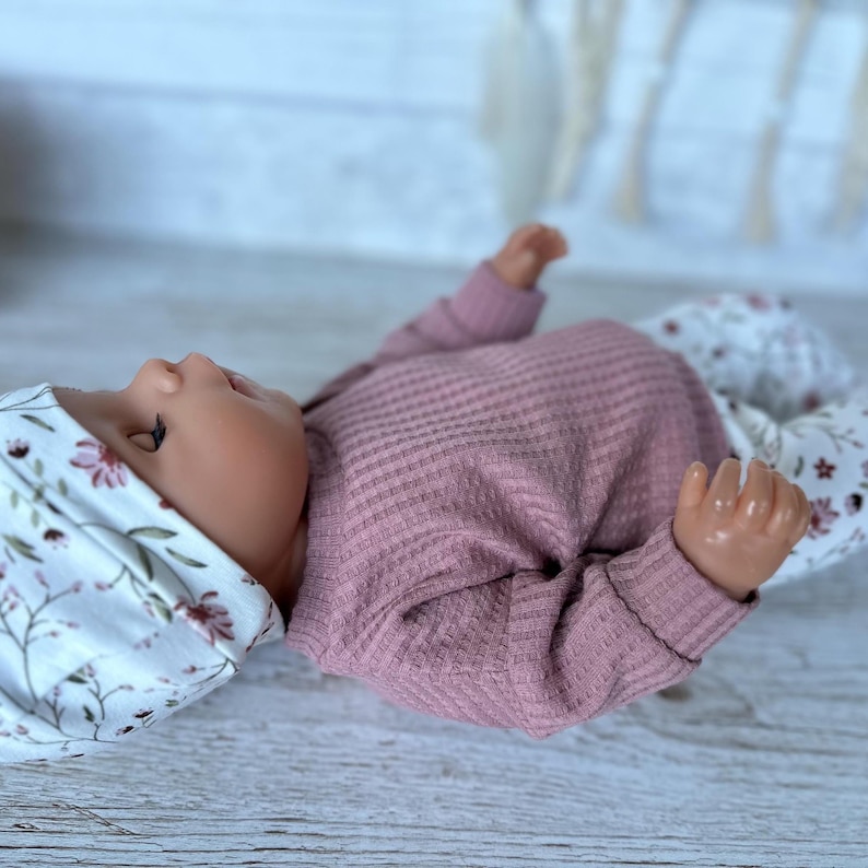 May include: A doll wearing a dusty rose long-sleeved waffle knit shirt, white pants with a floral pattern of small brown and pink flowers, and a matching white floral beanie. The doll has a smooth, light-toned complexion and is lying on its side.