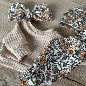 May include: A baby outfit set with a beige waffle knit top, floral print leggings, and a matching floral headband. The headband is tied in a bow.