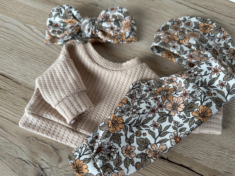 May include: A baby outfit set with a beige waffle knit top, floral print leggings, and a matching floral headband. The headband is tied in a bow.