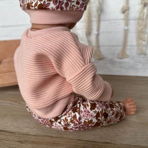 May include: A pink ribbed sweater with long sleeves and floral print trousers. The outfit is for a doll.