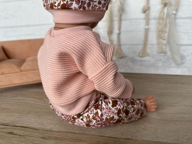 May include: A pink ribbed sweater with long sleeves and floral print trousers. The outfit is for a doll.