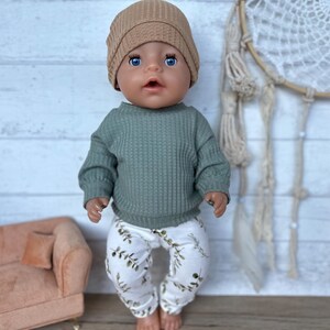 May include: A doll wearing a green waffle knit sweater, white pants with a green leaf pattern, and a brown beanie.