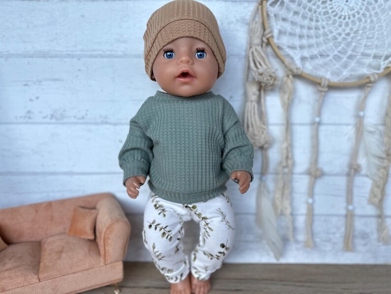 May include: A doll wearing a green waffle knit sweater, white pants with a green leaf pattern, and a brown beanie.