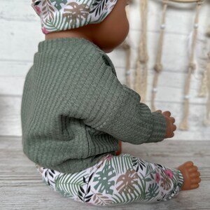 May include: A green ribbed sweater and floral print leggings for a doll. The leggings have a white background with green and brown leaves and pink flowers.