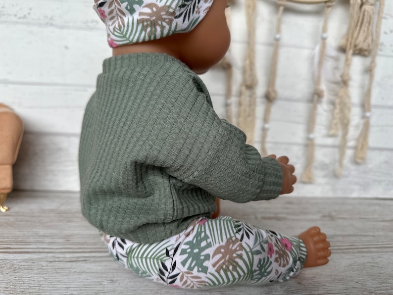 May include: A green ribbed sweater and floral print leggings for a doll. The leggings have a white background with green and brown leaves and pink flowers.