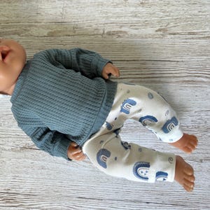 May include: A doll wearing a long-sleeved, slate blue waffle-knit shirt and off-white pants featuring a repeating pattern of small blue rainbows and swirls. The doll has a smooth, light-toned complexion and is positioned horizontally.