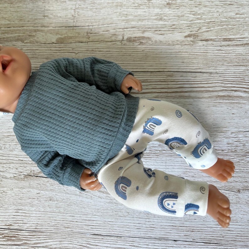 May include: A doll wearing a long-sleeved, slate blue waffle-knit shirt and off-white pants featuring a repeating pattern of small blue rainbows and swirls. The doll has a smooth, light-toned complexion and is positioned horizontally.