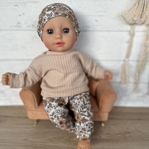 May include: A doll wearing a floral headband, a beige sweater, and floral pants sits on a small brown couch.