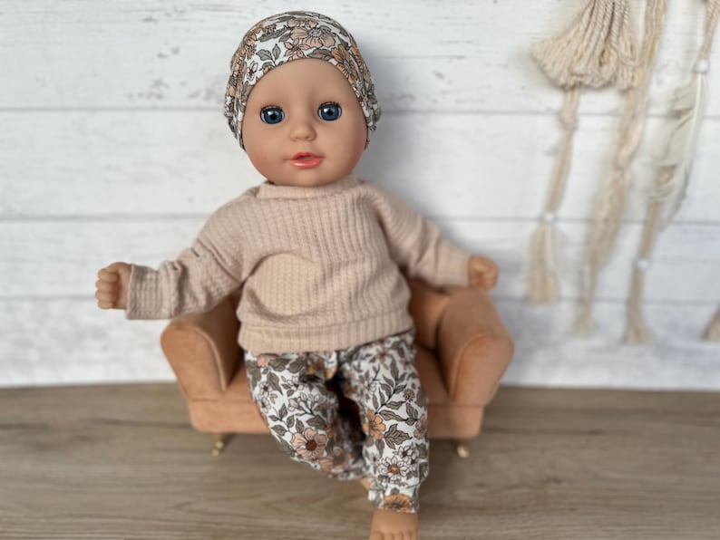May include: A doll wearing a floral headband, a beige sweater, and floral pants sits on a small brown couch.