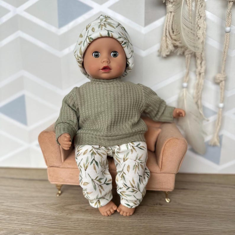 Pu&ograve; includere: Una bambola dalla pelle scura e occhi marroni indossa una maglia a maniche lunghe a coste color verde salvia, pantaloni bianchi con stampa di foglie verdi e marroni e un berretto abbinato, seduta su una piccola poltrona rosa pallido.