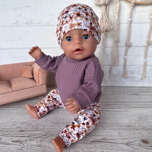 May include: A doll dressed in a purple waffle-knit top, floral trousers, and a matching hat. The doll is seated on a wooden surface. A small pink sofa is visible in the background.