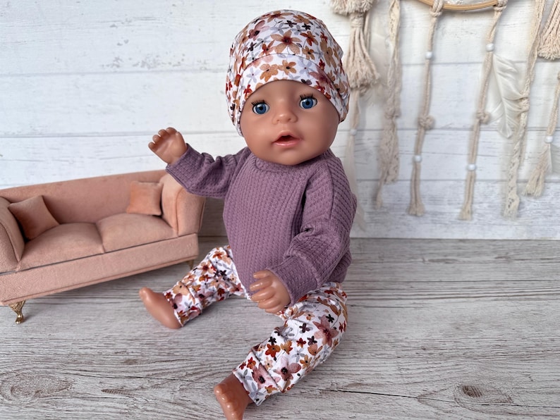 May include: A doll dressed in a purple waffle-knit top, floral trousers, and a matching hat. The doll is seated on a wooden surface. A small pink sofa is visible in the background.