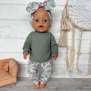 May include: A doll wearing a green waffle knit sweater and floral print pants with a matching floral headband. The doll is standing on a wooden surface with a dreamcatcher in the background.