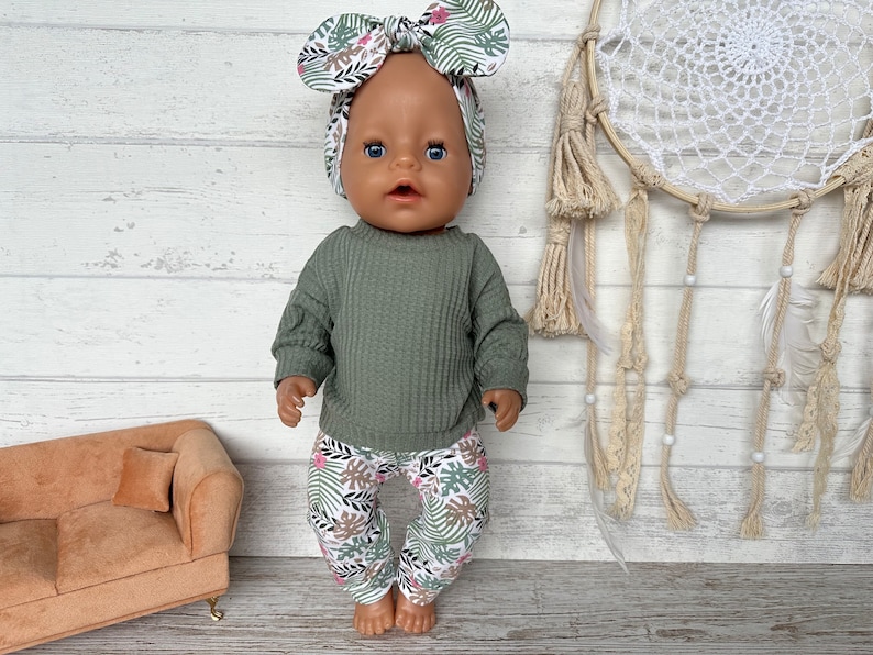 May include: A doll wearing a green waffle knit sweater and floral print pants with a matching floral headband. The doll is standing on a wooden surface with a dreamcatcher in the background.