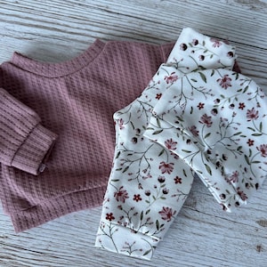 Puppenkleidung 43 cm 3-tlg Set bestehend aus einem mauvefarbenen, Langarm-Oberteil und einem passenden Set aus Hose und Mütze mit floralem Muster. Die Leggings und der Hut haben einen weißen Hintergrund mit einem Muster aus burgunderfarbenen Blumen und grünen Blättern.