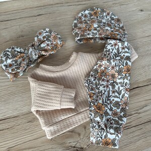 May include: A baby outfit set with a cream waffle knit top, floral print leggings, a matching headband, and a floral print beanie.