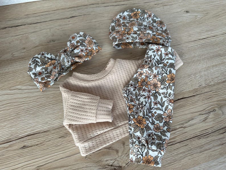 May include: A baby outfit set with a cream waffle knit top, floral print leggings, a matching headband, and a floral print beanie.
