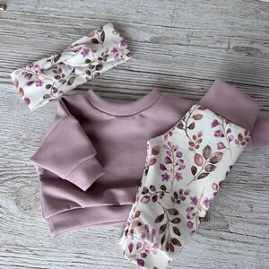 May include: A mauve long-sleeve sweatshirt, matching leggings with a white floral and leaf pattern, and a coordinating knotted headband. The leggings feature a repeating print of small purple flowers and brown leaves on a white background.