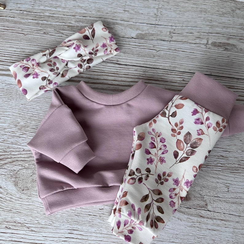 May include: A mauve long-sleeve sweatshirt, matching leggings with a white floral and leaf pattern, and a coordinating knotted headband. The leggings feature a repeating print of small purple flowers and brown leaves on a white background.