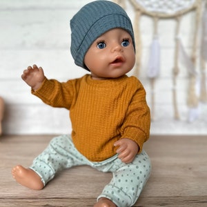 Può includere: Una bambola con occhi azzurri indossa un berretto blu in maglia a nido d'ape, una maglia a maniche lunghe color giallo senape in maglia a nido d'ape e pantaloni verde chiaro con un piccolo motivo a pois verde scuro. La bambola è seduta.
