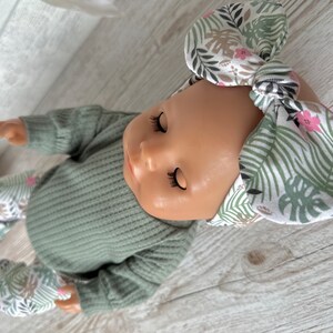 May include: A doll wearing a green ribbed sweater and matching pants with a white and green floral print. The doll is wearing a white headband with a green and pink floral print.