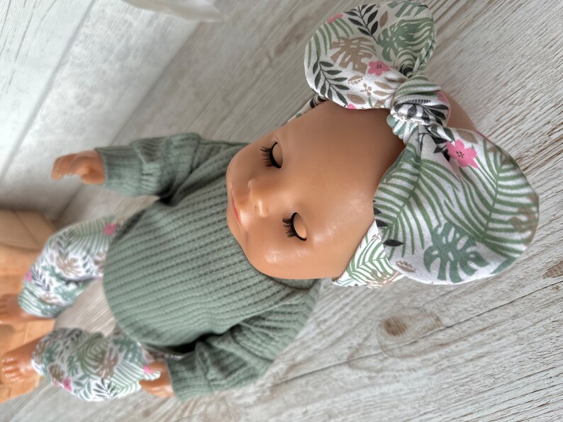 May include: A doll wearing a green ribbed sweater and matching pants with a white and green floral print. The doll is wearing a white headband with a green and pink floral print.