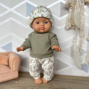 Pu&ograve; includere: Una bambola con viso rotondo e occhi scuri, che indossa una maglia a maniche lunghe a coste color verde oliva, pantaloni bianchi con stampa di foglie verdi e un berretto bianco abbinato con la stessa stampa di foglie verdi.