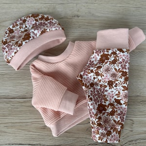 May include: A pink baby outfit set with a floral print. The set includes a long-sleeved sweater, trousers, and a hat.