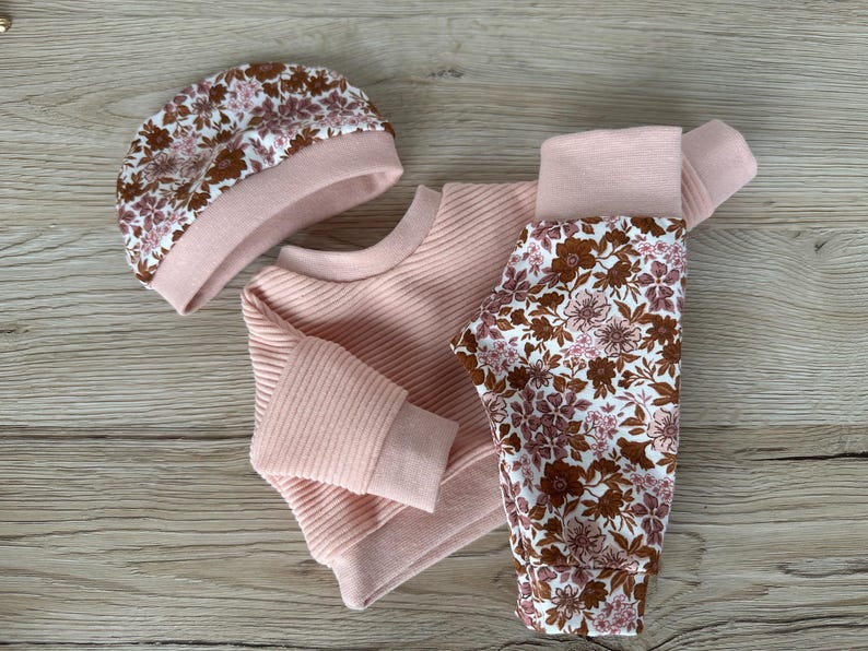 May include: A pink baby outfit set with a floral print. The set includes a long-sleeved sweater, trousers, and a hat.