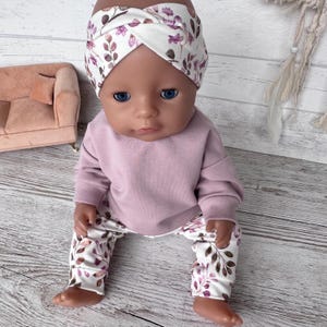 May include: A doll wearing a mauve long-sleeved sweatshirt and white leggings featuring a purple and brown leaf pattern. The doll also wears a matching white headband with a twisted front detail, decorated with the same purple and brown leaf print.