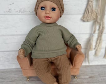 Pode incluir: Uma boneca com olhos azuis, vestindo um gorro de malha marrom, um suéter verde e calças marrons. A boneca está sentada em uma pequena cadeira cor de pêssego. A roupa da boneca tem uma textura canelada.