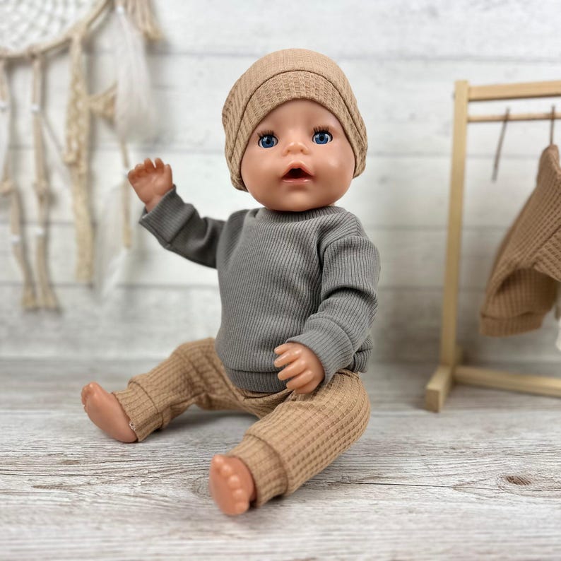 May include: A doll with blue eyes wearing a tan waffle-knit beanie, a long-sleeved grey ribbed shirt, and matching tan waffle-knit trousers. The doll is seated in a neutral pose, showcasing the textured fabric of the clothing set in a clear, detailed view.