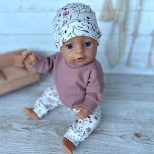 May include: A doll wearing a white beanie with a floral pattern, a long-sleeved mauve waffle-knit shirt, and matching white floral-patterned pants. The doll has blue eyes and is seated in a forward-facing position with its arms and legs extended.