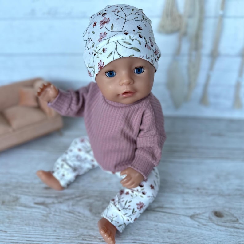 May include: A doll wearing a white beanie with a floral pattern, a long-sleeved mauve waffle-knit shirt, and matching white floral-patterned pants. The doll has blue eyes and is seated in a forward-facing position with its arms and legs extended.