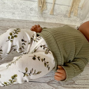 K&ouml;nnte beinhalten: Eine Babypuppe tr&auml;gt ein gr&uuml;nes Waffelstrick-Langarmshirt und eine wei&szlig;e Hose mit gr&uuml;nem Bl&auml;tterdruck.