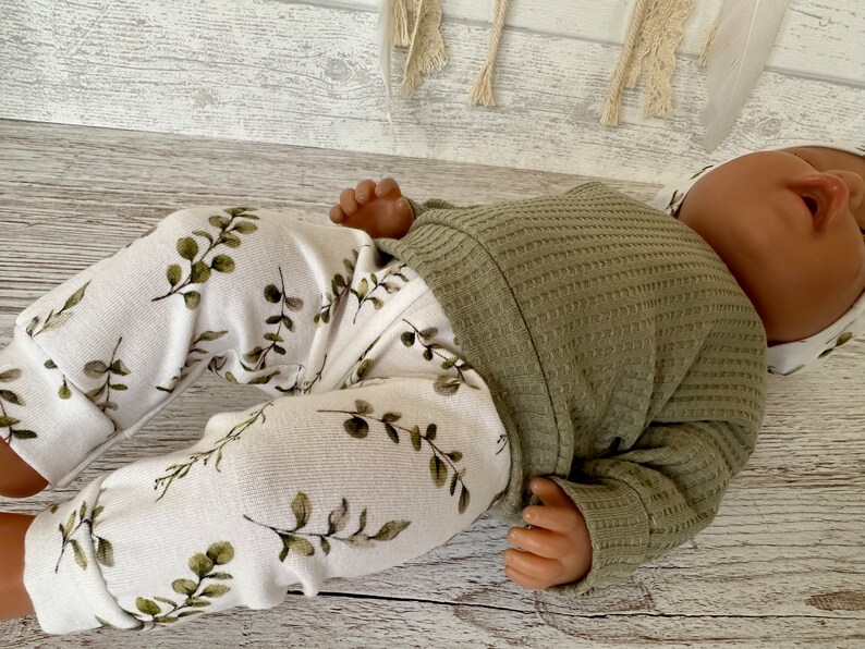 K&ouml;nnte beinhalten: Eine Babypuppe tr&auml;gt ein gr&uuml;nes Waffelstrick-Langarmshirt und eine wei&szlig;e Hose mit gr&uuml;nem Bl&auml;tterdruck.