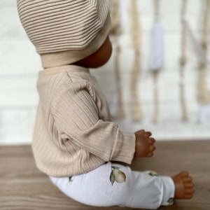 Puede incluir: Un top de punto de canal&eacute; beige y leggings blancos con estampado de lim&oacute;n. El atuendo lo lleva una mu&ntilde;eca con un gorro de punto beige.