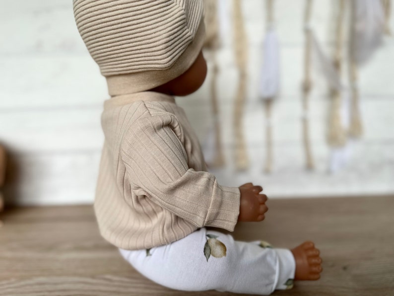 Puede incluir: Un top de punto de canal&eacute; beige y leggings blancos con estampado de lim&oacute;n. El atuendo lo lleva una mu&ntilde;eca con un gorro de punto beige.
