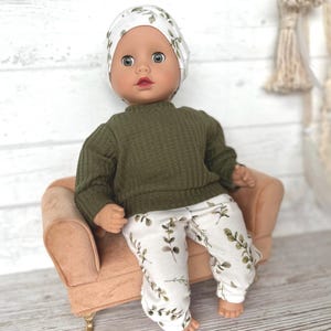 May include: A doll wearing a green sweater and white trousers with a leafy pattern. The doll is wearing a white and green patterned hat and is sitting on a small, light brown sofa.