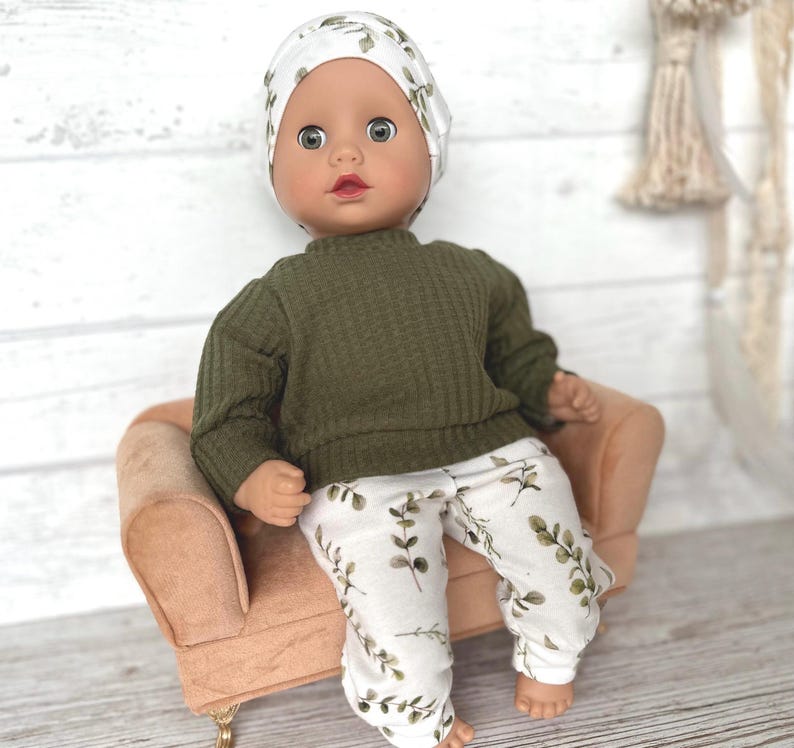 May include: A doll wearing a green sweater and white trousers with a leafy pattern. The doll is wearing a white and green patterned hat and is sitting on a small, light brown sofa.