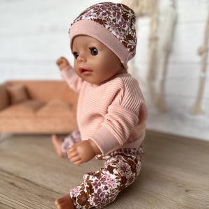 May include: A doll wearing a pink and brown floral print beanie, a pink ribbed sweater, and floral print trousers.