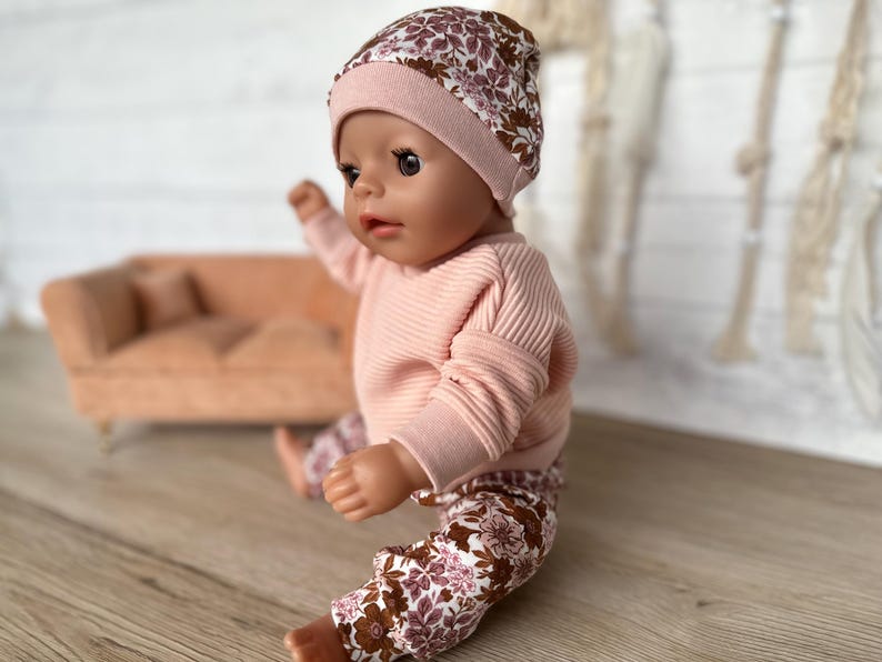 May include: A doll wearing a pink and brown floral print beanie, a pink ribbed sweater, and floral print trousers.