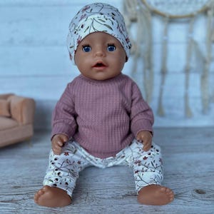 May include: A doll with blue eyes wearing a long-sleeved mauve ribbed shirt and white pants featuring a delicate floral pattern of small red flowers and green leaves. The doll also wears a matching white beanie with the same floral print design.