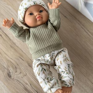 Pu&ograve; includere: Una bambola con occhi azzurri indossa una maglia a maniche lunghe in tessuto a nido d'ape color verde salvia, pantaloni bianchi con motivo a foglie verdi e marroni e un berretto abbinato. La bambola ha le braccia sollevate e indossa abiti coordinati.