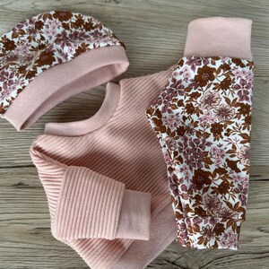 May include: A pink baby outfit set with a floral print. The set includes a long-sleeved sweater, leggings, and a beanie.