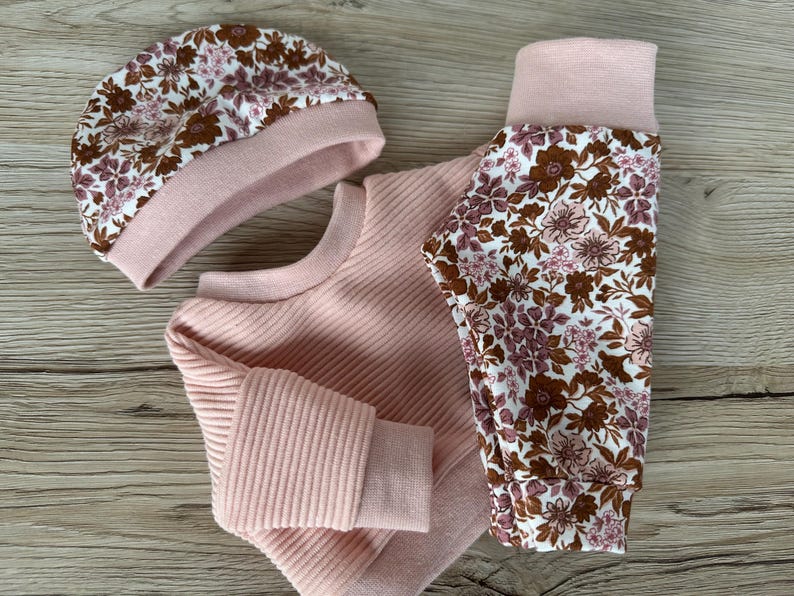 May include: A pink baby outfit set with a floral print. The set includes a long-sleeved sweater, leggings, and a beanie.