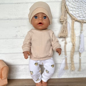 Puede incluir: Una mu&ntilde;eca beige que lleva un su&eacute;ter beige de canal&eacute; y pantalones blancos con estampado de lim&oacute;n. La mu&ntilde;eca lleva una boina beige. La mu&ntilde;eca est&aacute; de pie frente a un fondo de madera blanco.