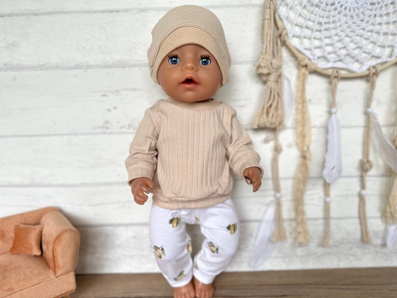 Puede incluir: Una mu&ntilde;eca beige que lleva un su&eacute;ter beige de canal&eacute; y pantalones blancos con estampado de lim&oacute;n. La mu&ntilde;eca lleva una boina beige. La mu&ntilde;eca est&aacute; de pie frente a un fondo de madera blanco.