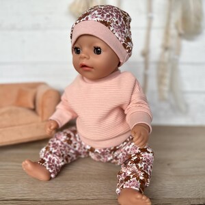 May include: A doll wearing a pink sweater and floral print trousers with a pink and floral print beanie.