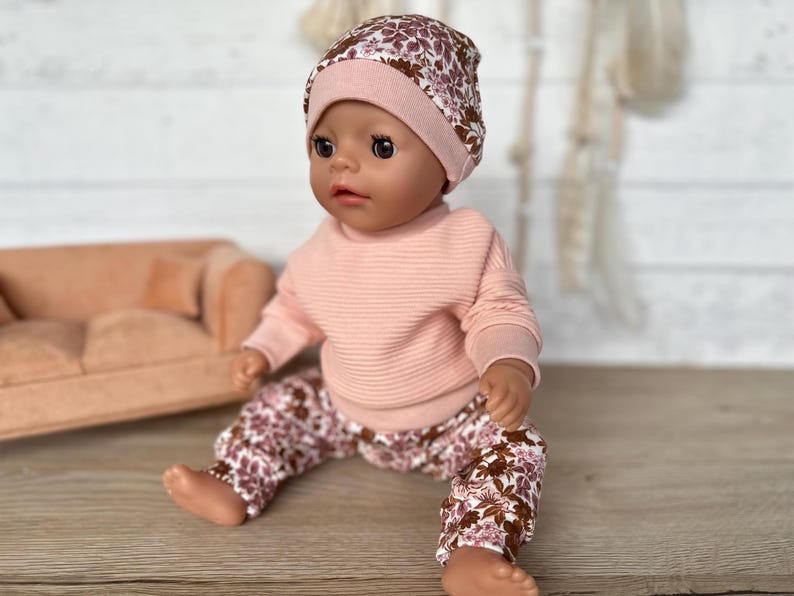May include: A doll wearing a pink sweater and floral print trousers with a pink and floral print beanie.